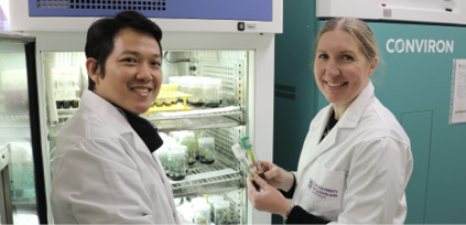 Two scientists in white lab coats stand next to a laboratory refrigerator. One is a man and one is a woman. They are both smiling. 