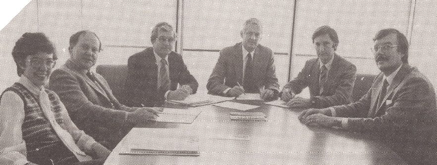 A black-and-white photo of five men in suits and one woman sitting around a board table. Their hands are on the table and they are holding pens, and there is paper scattered across the table.