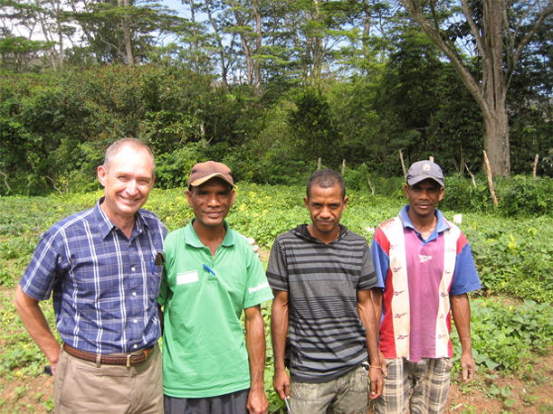 Four men standing in front of a field. There are trees in the background. Two men are wearing caps and they are all smiling. 