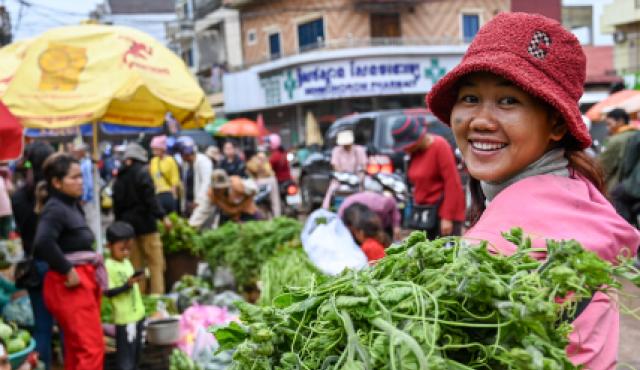 A woman at an outdoors busy market smiles at the camera while she holds a large bunch of green vegetables.