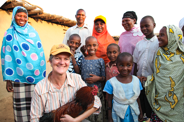 A woman in a yellow cap kneels at the front holding a chicken. Behind her are a group of men, women and children who are smiling at the camera. One of the women is also wearing a yellow cap. Behind them is a small building with a thatch roof. 