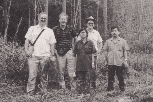A black-and-white photo of five people standing in front of a forest. One man is wearing a brimmed hat. They are all wearing long pants and short-sleeved shirts. 